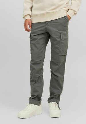Pantalons cargo en vert olive, fabriqués en coton résistant, avec des poches latérales et un design de jambe fuselée. Portés avec des baskets blanches.