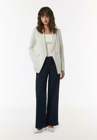 Biały żakiet z pojedynczym guzikiem, zestawiony z ciemnymi, szerokimi dżinsami i szarymi czółenkami na wysokim obcasie. Modelka ma kręcone włosy, minimalna biżuteria.