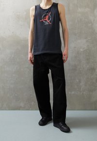 Mörkgrå ärmlös tanktopp med röd grafisk design, kombinerad med lösa svarta byxor och svarta snörskor, mot en strukturerad grå bakgrund.