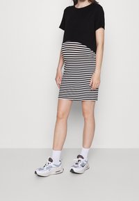 Haut noir à manches courtes fusionnant avec une jupe moulante rayée noire et blanche. Le mannequin porte des baskets blanches et bleues avec des accents gris.