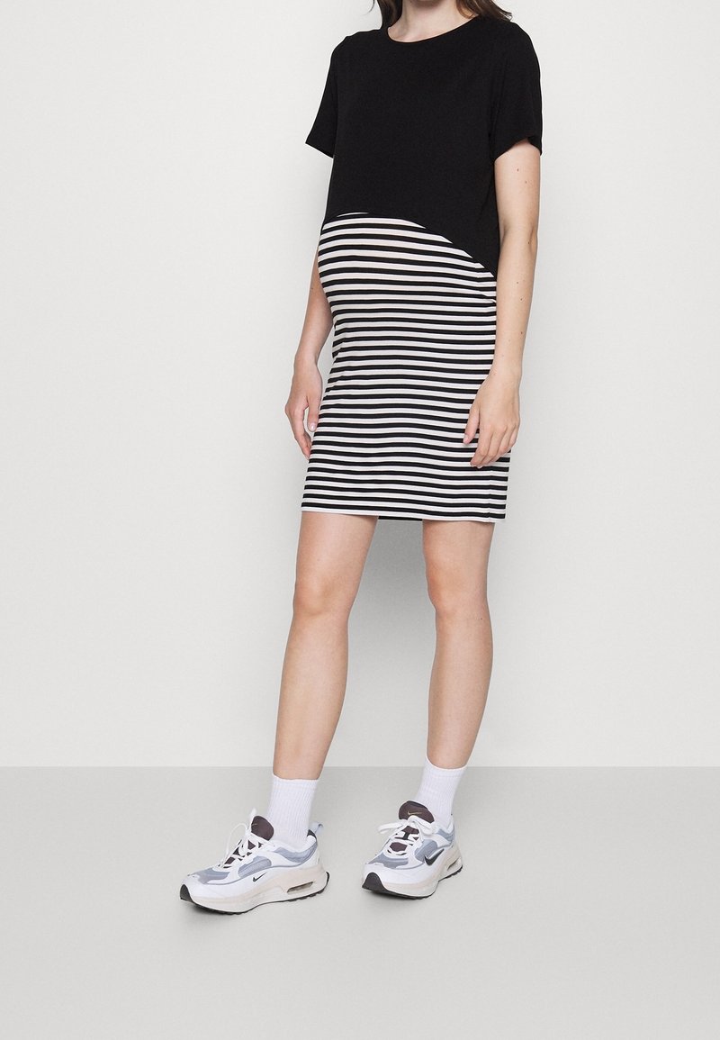 Haut noir à manches courtes fusionnant avec une jupe moulante rayée noire et blanche. Le mannequin porte des baskets blanches et bleues avec des accents gris.