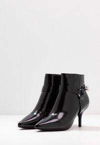 Botines de charol negro con punta afilada y un diseño elegante. Presenta una correa decorativa y acentos metálicos en el lateral.