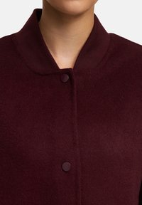 Bourgondische wollen jas met een gebreide kraag, voorzien van een frontsluiting met knopen en een zachte textuur. Focus op minimalistische ontwerpelementen.