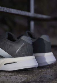 Par sorte og grå løbesko mærket "Adizero Boston 13" på en tekstureret udendørs overflade.