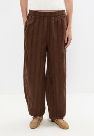 Femme portant un pantalon marron ample avec des poches latérales et des chaussures plates beiges, debout devant un fond blanc uni.
