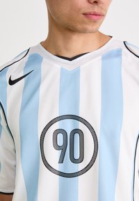 Hvit og lyseblå stripete trøye med svarte detaljer, med en sirkulær svart grafikk med "90" i midten. V-hals design.