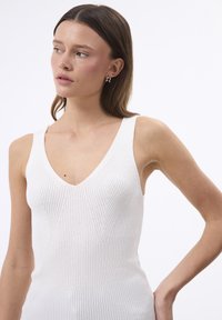 Valge ribakudumiga tank-top sügava V-kaelusega, kitsas sobivus ja tekstuuriga vertikaalsed jooned. Mudelil on pikad juuksed ja lihtsad kõrvarõngad.