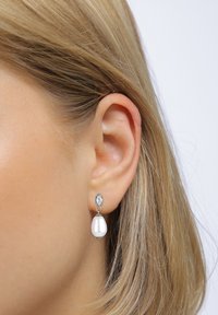 Die Ohrringe hängen in einem silberfarbenen Setting mit klaren Edelsteinen und einem schimmernden weißen Perlen-Tropfen-Design.