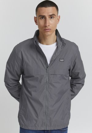 Graue Zip-Jacke mit Stehkragen und kleinem Logo auf der Brust. Merkmale sind eine leichte, glatte Textur und elastische Bündchen.