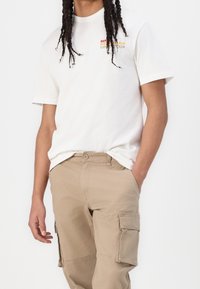 Weißes Baumwoll-T-Shirt mit mehrfarbigem "Monte Carlo"-Schriftzug. Die Person trägt khakifarbene Cargo-Hosen mit seitlichen Taschen und einer taillierten Passform.