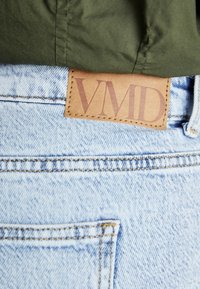 Heledad teksad helesinises värvitoonis, millel on nahklabel, millele on reljeefselt trükitud "VMD", ning kontrastne õmblus vöökoha ja taskute ääres.