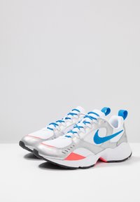 Baskets Nike avec une tige synthétique argentée, un mesh blanc, des lacets et des accents bleus, présentant un détail de talon rose et une semelle noire.