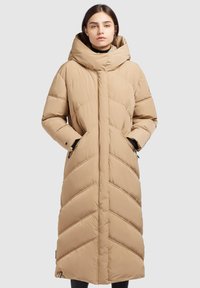 Manteau doudoune beige avec un col haut, une fermeture éclair devant et des coutures en chevrons. Longueur longue avec des poches latérales et une texture lisse.