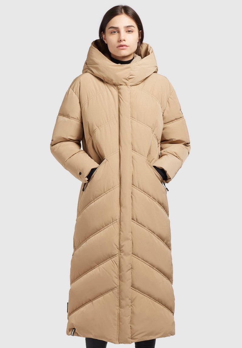Manteau doudoune beige avec un col haut, une fermeture éclair devant et des coutures en chevrons. Longueur longue avec des poches latérales et une texture lisse.