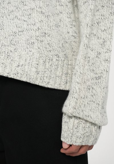 Lys grå strikkegenser med teksturfinish og ribbede mansjetter, kombinert med svarte bukser, som fremhever et avslappet design.