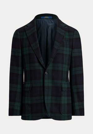 Homme portant des lunettes de soleil, un pull à col roulé en maille torsadée bleu marine, un blazer à carreaux foncé, un jean usé, une ceinture marron et des mocassins à pampilles marron, debout les mains dans les poches.
