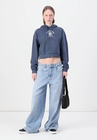 Beskjært marineblå hettegenser med frontlomme og hvit logo, kombinert med lyseblå vide jeans. Svart veske med stropp og svarte sneakers.