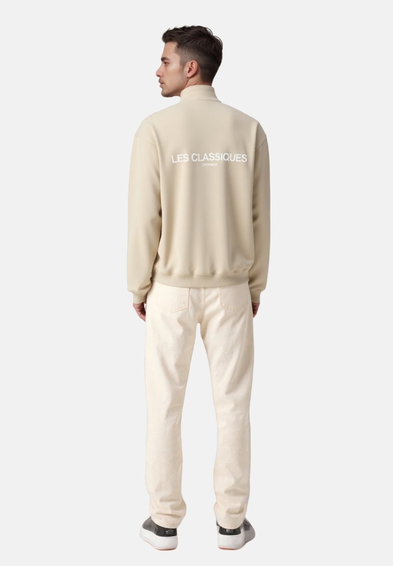Giacca in pile beige con colletto alto e testo bianco "LES CLASSIQUES" sul retro, abbinata a pantaloni chiari e sneakers bianche.