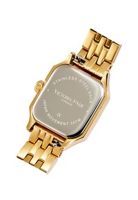 Montre en acier inoxydable doré avec un boîtier rectangulaire, un bracelet métallique et un fond argenté réfléchissant portant un logo gravé et des détails.