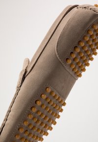 Beige suede slip-on shoe with rubberized, rounded accents along one side. Features a textured surface and minimal stitching detail.