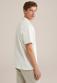 Camicia a maniche corte con colletto in tessuto bianco chiaro con un motivo testurizzato, abbinata a pantaloni beige chiaro, indossata da un modello di profilo.