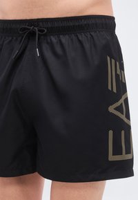Μαύρο μαγιό βερμούδα με ελαστική μέση, κορδόνι και χρυσές διακεκομμένες λεπτομέρειες λογότυπου στο πλάι. Λείο, ελαφρύ ύφασμα.