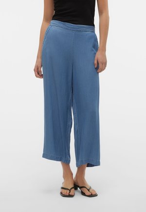 Vrouw draagt losse, wijde blauwe cropped broek gecombineerd met een zwart mouwloos topje en zwarte sandalen met open tenen tegen een witte achtergrond.