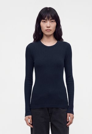 Femme aux longs cheveux noirs portant un haut bleu marine ajusté, côtelé et à manches longues, ainsi qu'un pantalon noir, debout devant un fond blanc uni.