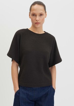 Donna con i capelli tirati indietro indossa un maglione nero ampio a maniche corte e pantaloni di jeans blu, in piedi con le mani nelle tasche davanti a uno sfondo semplice.