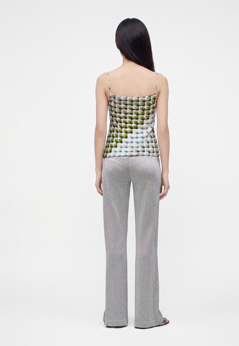 Femme aux longs cheveux foncés portant un haut sans manches à motifs et un pantalon large gris clair, debout de dos sur un fond uni.