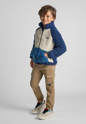 Fleecejacke in Creme, Marineblau und Blau mit hohem Kragen, Reißverschluss, Vorderentaschen und einer weichen Textur. Kombiniert mit beigen Cargo-Hosen und Turnschuhen.