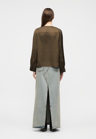 Γυναίκα με μακριά καστανά μαλλιά που φοράει καφέ μπλούζα με μακριά μανίκια και φούστα maxi από ανοιχτό μπλε τζιν με σχίσιμο πίσω και μαύρα παπούτσια.