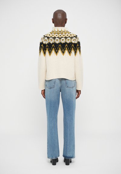 Persona che indossa un maglione a maglia color crema con un motivo geometrico in nero e giallo sul colletto, jeans azzurri chiari e stivali neri con tacco, in piedi di spalle.
