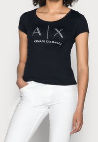 Černé bavlněné tričko s krátkými rukávy se zdobeným logem "A|X ARMANI EXCHANGE" na přední straně, v kombinaci s bílými slim-fit džíny.