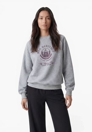 Kvinde med langt mørkt hår iført en grå sweatshirt med lilla Neo Heritage-logo og sorte bukser, stående mod en hvid baggrund.