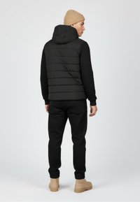 Black puffer vest with horizontal stitching, worn over a black sweatshirt, paired with black pants and tan boots; features a hood and casual fit.