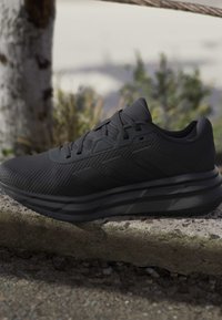 Baskets sportives noires avec tige texturée et semelle épaisse posées sur un rebord en pierre en extérieur avec un arrière-plan de verdure flou.