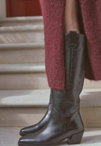 Botas de cuero negras hasta la rodilla con punta cuadrada y tacón grueso, combinadas con un abrigo de lana texturizada burdeos. Sentada en escalones de piedra.