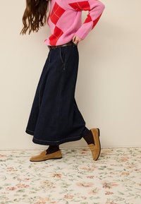 Personne portant un pull irlandais rose et rouge avec une jupe trapèze en jean foncé et des mocassins marron sur un tapis à motifs floraux.