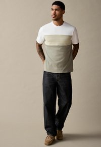 Kortærmet farveblokeret t-shirt med en hvid øverste sektion, textureret lys grøn midte og mørkere grøn bund, parret med mørke denimbukser.