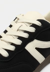 Μαύρο αθλητικό παπούτσι με κρεμ κορδόνια και ανάγλυφο λευκό λογότυπο στο πλάι. Το επάνω μέρος αποτελείται από μίγμα λείου και σουέντ υλικού. Λαστιχένια σόλα.