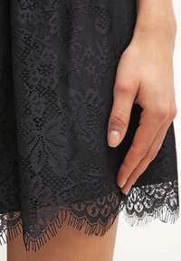 Black lace dress detail featuring floral patterns, a textured surface, and a scalloped hem with fringed accents. Hand gently resting on skirt.