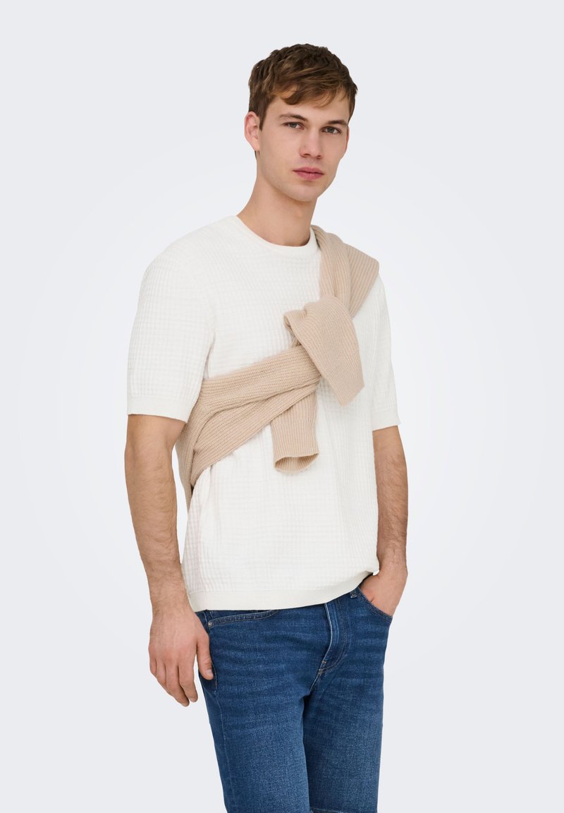 Jeune homme portant une chemise blanche texturée à manches courtes et un jean bleu, avec un pull beige clair drapé sur ses épaules.