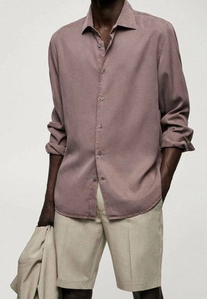 Mand iført en violet langærmet skjorte med opkrogede ærmer, beige shorts, holder en beige jakke i venstre hånd.