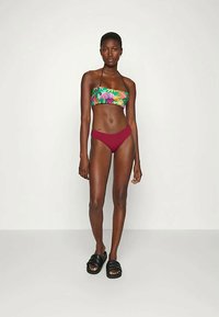 Mulher de pé, usando um top de bikini bandeau floral, parte inferior de bikini marrom sólido e sandálias slides pretas, contra um fundo branco liso.