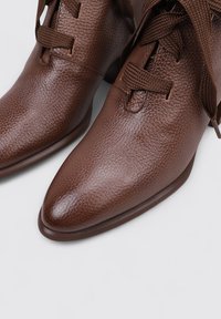 Botas de tobillo de cuero marrón con un acabado texturizado, puntera afilada y detalle de cordones texturizados. Suela lisa con un tacón bajo.
