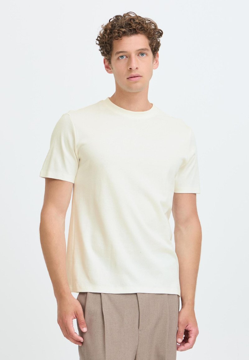 Νέος με σγουρά μαλλιά, φορώντας μια απλή off-white κοντομάνικη μπλούζα και μπεζ παντελόνι, στέκεται μπροστά σε ένα λευκό φόντο.
