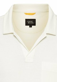 Hvid polo shirt med et struktureret mønster, flad krave, en brystlomme og en sort label med "camel active" og størrelse "L."