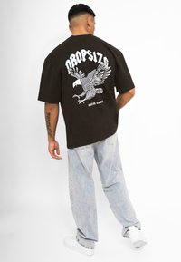Brown oversized t-shirt with a white eagle graphic and text on the back, paired with light blue baggy jeans and white sneakers.