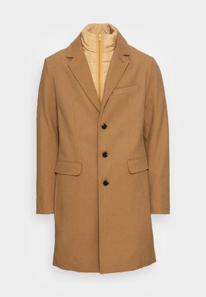Manteau long en laine couleur chameau avec revers, boutons noirs, deux poches à rabat et une couche intérieure matelassée beige avec fermeture éclair visible au niveau du col.